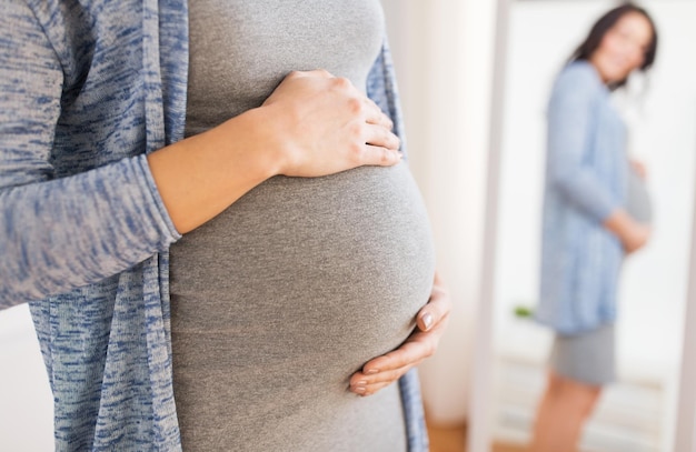 concepto de embarazo, maternidad, personas y expectativas: cerca de una mujer embarazada feliz con una gran barriga mirando al espejo en casa