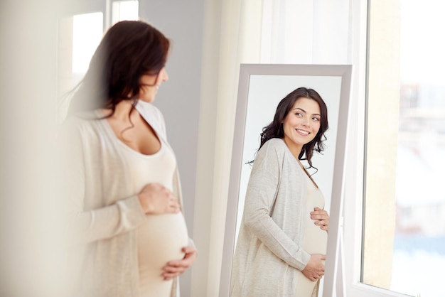 concepto de embarazo, maternidad, personas y expectativas - mujer embarazada feliz mirando al espejo en casa