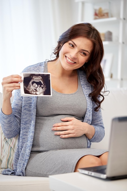 concepto de embarazo, maternidad, personas y medicina - mujer embarazada feliz con computadora portátil que tiene una videollamada y muestra una imagen de ultrasonido en casa