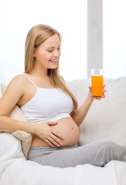 concepto de embarazo, maternidad, salud, comida y felicidad - mujer embarazada feliz sentada en un sofá con jugo de naranja fresco