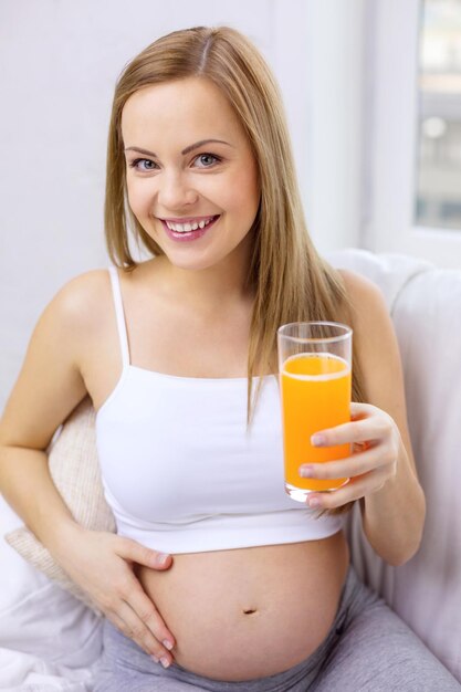 concepto de embarazo, maternidad, salud, comida y felicidad - mujer embarazada feliz sentada en un sofá con jugo de naranja fresco