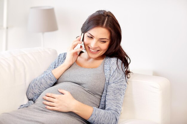 concepto de embarazo, maternidad, tecnología, personas y expectativas - mujer embarazada feliz llamando al teléfono inteligente en casa