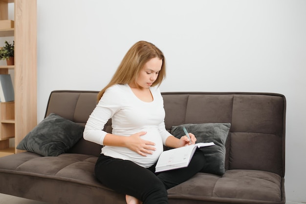 Concepto de embarazo, personas y expectativa - mujer embarazada feliz escribiendo en el cuaderno en casa