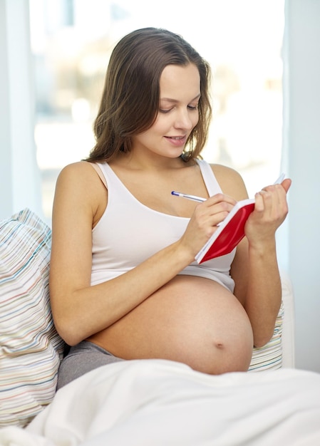 Concepto de embarazo, personas y expectativa - mujer embarazada feliz escribiendo en el cuaderno en casa