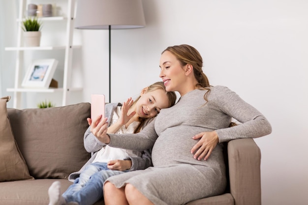 concepto de embarazo, personas y familia - feliz mujer embarazada y niña sentada en el sofá y tomando selfie por teléfono inteligente en casa