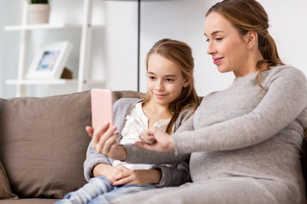 concepto de embarazo, personas y familia - feliz mujer embarazada y niña sentada en el sofá y tomando selfie por teléfono inteligente en casa