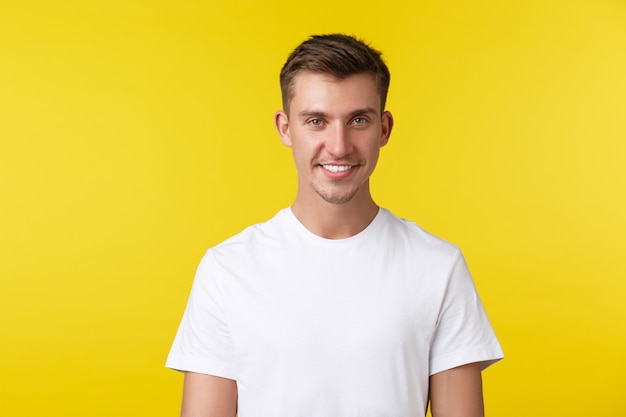 Concepto de emociones de estilo de vida, verano y personas. Retrato de primer plano de un joven apuesto en camiseta blanca casual, sonriendo a la cámara complacido, de pie fondo amarillo.