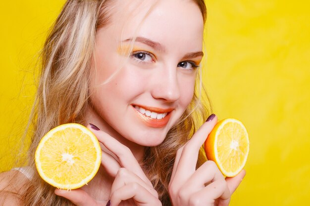 Concepto de emociones, salud, personas, comida y belleza - Beauty Model Girl toma Juicy Oranges. Hermosa chica adolescente alegre con pecas, peinado divertido. maquillaje amarillo. Maquillaje profesional. Rodaja de naranja