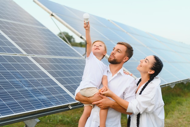 Concepto de energía solar Una joven familia feliz está parada cerca de paneles solares y sosteniendo una bombilla eléctrica