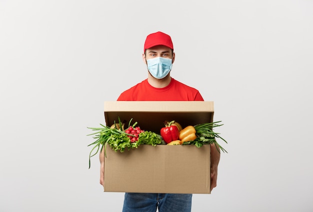 Concepto de entrega: apuesto hombre de mensajería de entrega de comestibles caucásico en uniforme rojo y mascarilla con caja de supermercado con frutas y verduras frescas