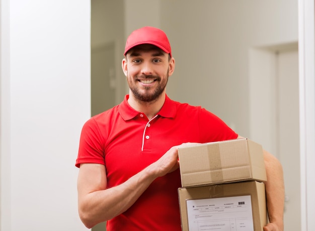 concepto de entrega, correo, personas y envío - hombre feliz con uniforme rojo con cajas de paquetes en el pasillo