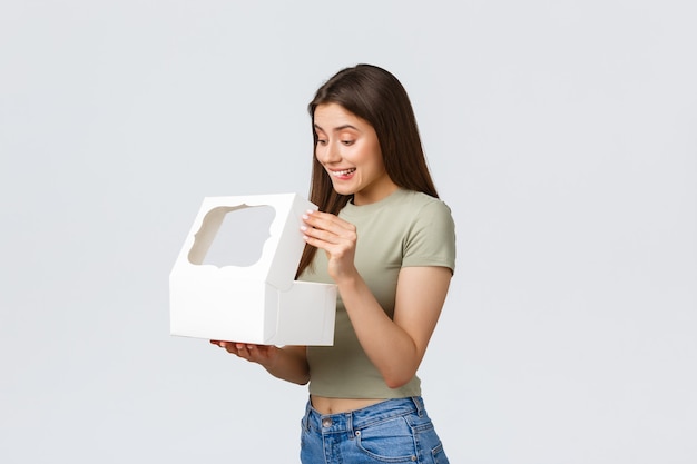 Concepto de entrega, estilo de vida y comida. Sonriente novia feliz abrir caja blanca con dulces y deliciosos postres, pasteles o muffins ordenados en la mejor pastelería, de pie fondo blanco.