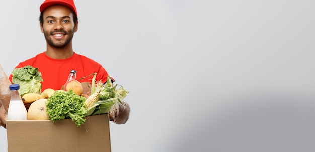 Concepto de entrega Guapo repartidor afroamericano que lleva una caja de comida y bebida de la tienda aislada en el espacio de copia de fondo del estudio gris