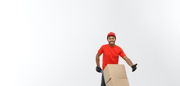 Concepto de entrega Retrato de un apuesto repartidor afroamericano o un mensajero empujando un camión de mano con una pila de cajas aisladas en el espacio de copia de fondo del estudio gris