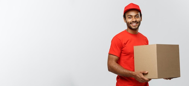 Concepto de entrega Retrato del repartidor afroamericano feliz en tela roja sosteniendo un paquete de caja aislado en el espacio de copia de fondo de estudio gris