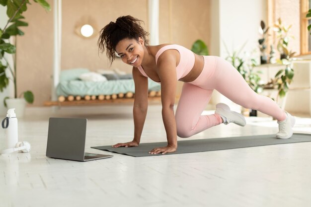 Concepto de entrenamiento doméstico Dama negra haciendo ejercicio de alpinistas de cuerpo cruzado viendo tutorial en línea en una computadora portátil