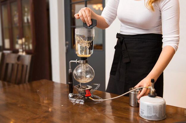 concepto de equipo, cafetería, personas y tecnología - cierre de una mujer con quemador de gas butano calentando agua en una cafetera de sifón en el bar cafetería o en la cocina del restaurante