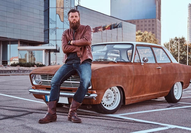 Concepto de estilo campestre. Hombre barbudo vestido con chaqueta de cuero marrón y botas apoyado en un auto retro sintonizado en el estacionamiento de la ciudad cerca de un rascacielos.