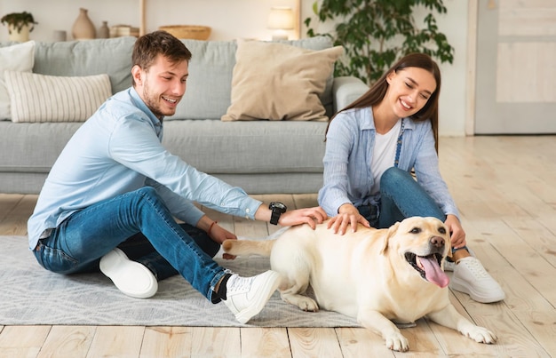 Concepto de estilo de vida acogedor. Feliz pareja joven rascarse su adorable perro labrador sentados en el suelo en la moderna sala de estar. Retrato, de, sonriente, hombre y mujer, juego, con, perro, amigo, adentro