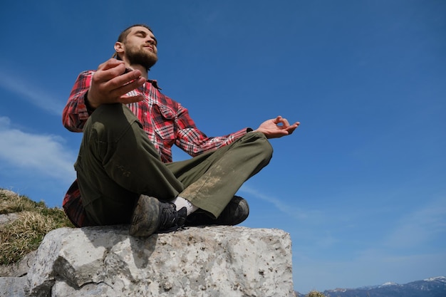 Concepto de estilo de vida activo Joven con rastas medita y hace yoga afuera contra la vista del cielo azul desde abajo Chico viajero sentado en una gran roca en las montañas y disfrutando de las vistas de la naturaleza