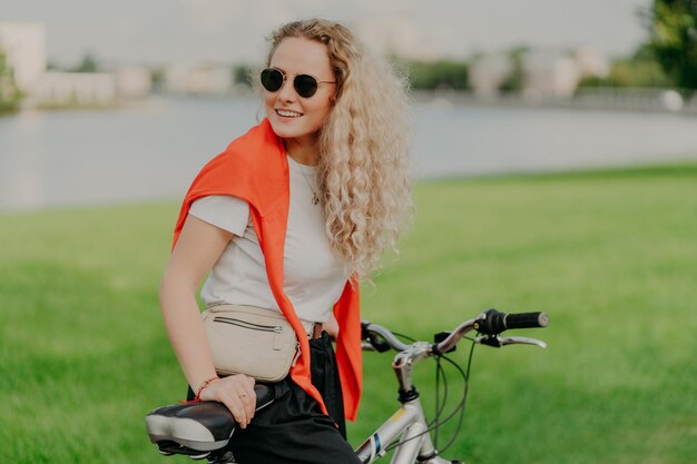 Concepto de estilo de vida activo Una mujer joven positiva con cabello rizado y claro monta en bicicleta en el césped verde usa una camiseta blanca y un suéter rojo en los hombros lleva una pequeña bolsa de buen humor