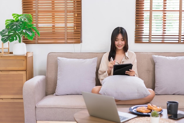 Concepto de estilo de vida en casa Mujer asiática joven escribiendo en tableta para trabajar mientras está sentada en el sofá