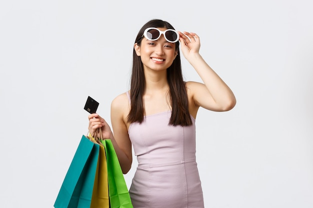 Concepto de estilo de vida, compras y turismo. Feliz mujer bonita asiática en vestido y gafas de sol, cámara sonriente