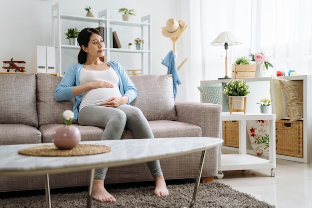 concepto de estilo de vida de cuidado de maternidad feliz. hermosa y elegante niña prepárese para mamá esperando un niño boca abajo sosteniendo el vientre relajándose en el sofá del sofá en un luminoso y acogedor apartamento moderno cuidado espera marido