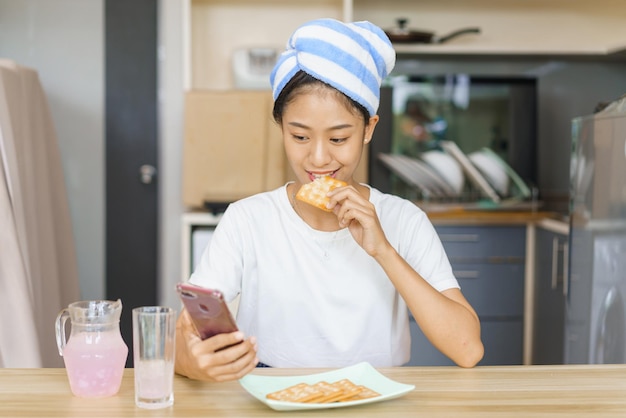 Concepto de estilo de vida doméstico Mujer joven en turbante después de la ducha para comer galletas y navegar en las redes sociales