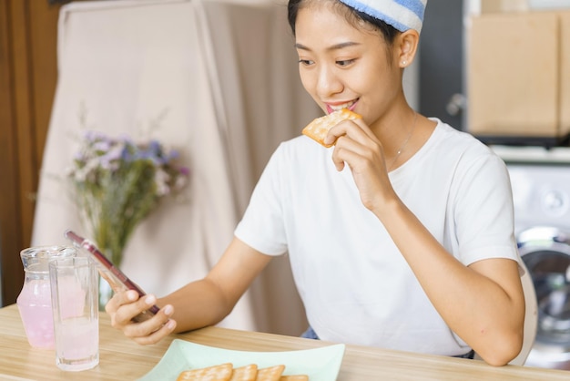 Concepto de estilo de vida doméstico Mujer joven en turbante después de la ducha para comer galletas y navegar en las redes sociales