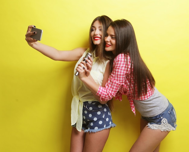 Concepto de estilo de vida, felicidad, emocional y personas: dos chicas hipster de belleza con un micrófono toman selfie sobre fondo amarillo