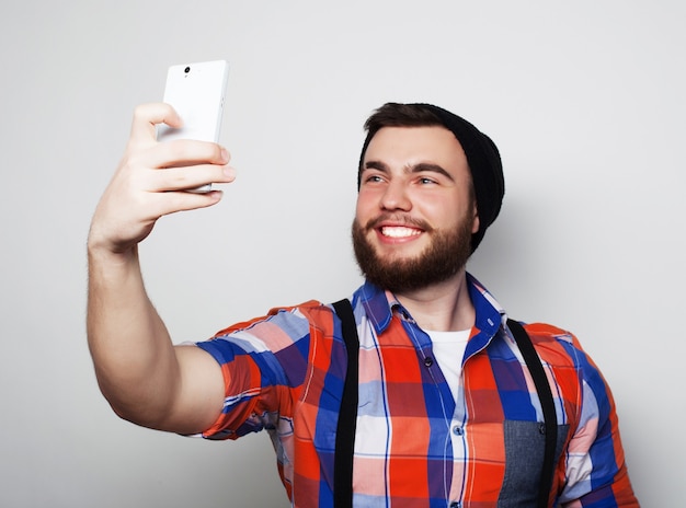 Concepto de estilo de vida. Un hombre joven con barba en camisa sosteniendo un teléfono móvil y haciendo una foto de sí mismo mientras está de pie en gris