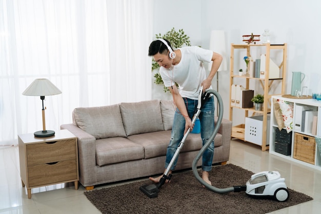 concepto de estilo de vida de limpieza del hogar masculino. un joven chino asiático con auriculares con aspiradora en la sala de estar de su casa arregla la alfombra. un chico guapo disfruta de la música en los auriculares mientras hace las tareas del hogar.