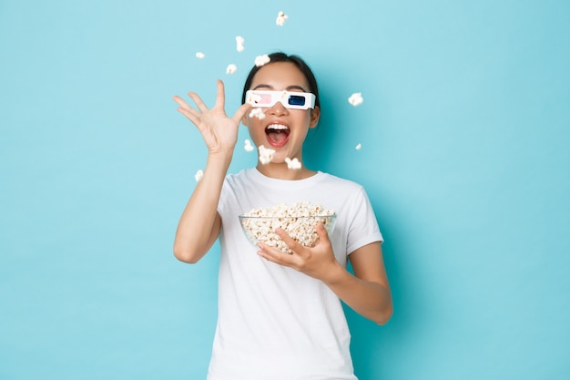 Concepto de estilo de vida, ocio y emociones. Impresionada y emocionada chica asiática disfrutando viendo películas, lanzando palomitas de maíz a la pantalla del televisor con gafas 3d, pared azul claro de pie