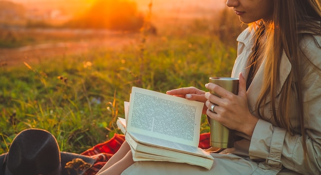 El concepto de estilo de vida y recreación familiar al aire libre en otoño. mujer con sombrero lee libros a cuadros con una taza térmica. Otoño. Puesta de sol. Acogedor