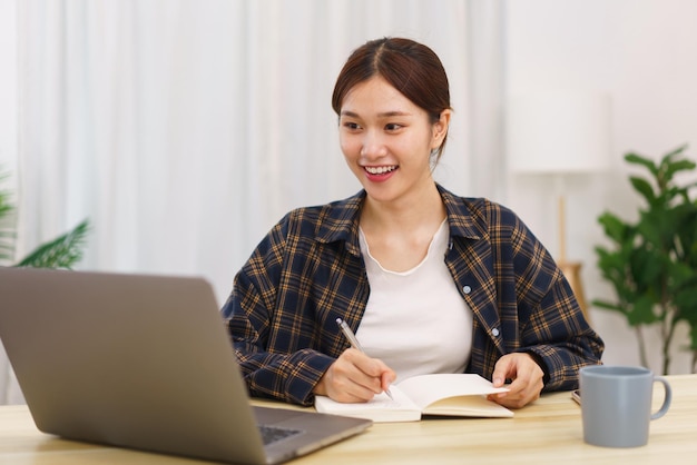 Concepto de estilo de vida en la sala de estar Mujer asiática joven que usa una computadora portátil y toma notas en el cuaderno