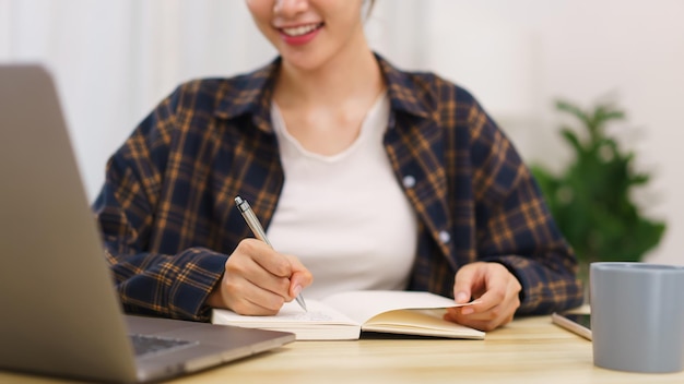 Concepto de estilo de vida en la sala de estar Mujer asiática joven que usa una computadora portátil y toma notas en el cuaderno