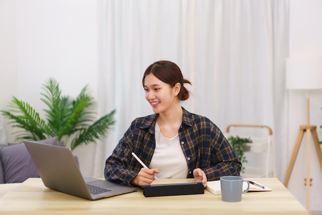 Concepto de estilo de vida en la sala de estar Mujer asiática joven que usa una computadora portátil y toma notas de datos en la tableta