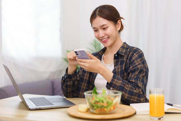 Concepto de estilo de vida en la sala de estar Mujer asiática joven que usa un teléfono inteligente y come ensalada de verduras