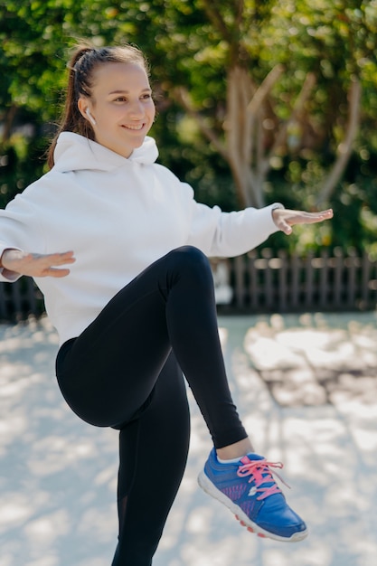 Concepto de estilo de vida saludable de deporte fitness. La mujer deportiva complacida trata de mantener el equilibrio sobre una pierna, mantiene la rodilla levantada, tiene entrenamiento a intervalos o entrenamiento cardiovascular, usa leggings blancos y zapatillas de deporte.