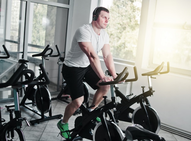 Concepto de estilo de vida saludable. Joven deportivo en camiseta blanca y pantalones cortos está ejercitando bicicleta en clase de spinning. Entrenamiento cardiovascular