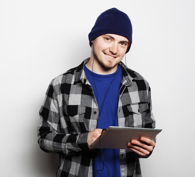 Concepto de estilo de vida, tecnología y personas: apuesto joven con camisa y sombrero trabajando en tableta digital y sonriendo mientras está de pie sobre fondo gris