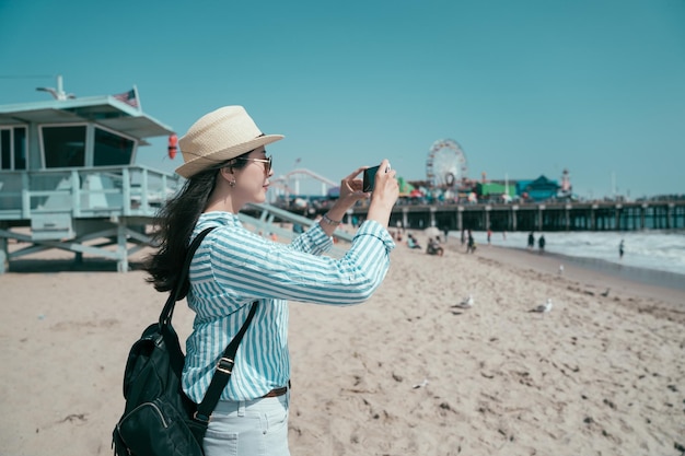 Concepto de estilo de vida de viaje. vista lateral hermosa turista asiática china con sombrero de paja y gafas de sol tomando fotos en el teléfono móvil mientras se encuentra en la playa de arena. chica viajera fotografiando celular