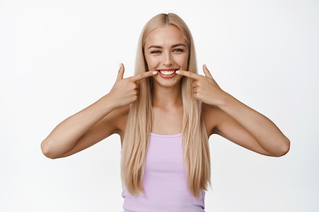 Concepto de estomatología y clínica dental Hermosa chica rubia mostrando su sonrisa blanca apuntando con el dedo a los dientes y con un fondo blanco satisfecho