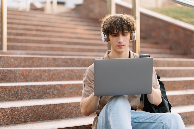 Concepto de estudio a distancia de elearning o concepto de aprendizaje remoto Joven feliz estudiante de colegio o estudiante universitario de pelo rizado con mochila y auriculares usando una computadora portátil sentado en los escalones de la universidad