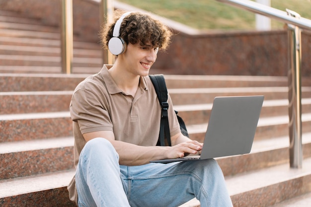 Concepto de estudio a distancia de elearning o concepto de aprendizaje remoto Joven feliz estudiante de colegio o estudiante universitario de pelo rizado con mochila y auriculares usando una computadora portátil sentado en los escalones de la universidad