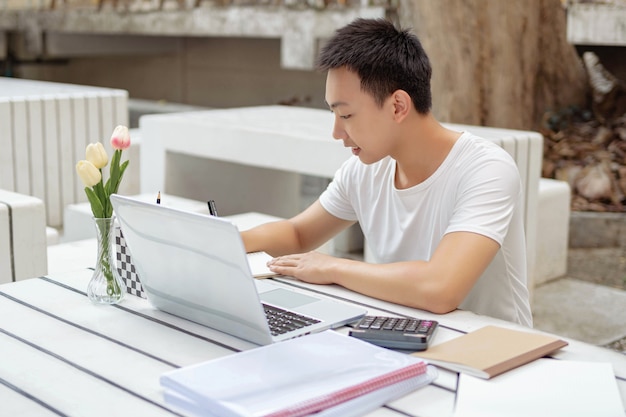 Concepto de estudio en línea un estudiante masculino en camiseta blanca disfrutando de estudiar en línea y sentado frente a su nueva computadora portátil blanca al aire libre.