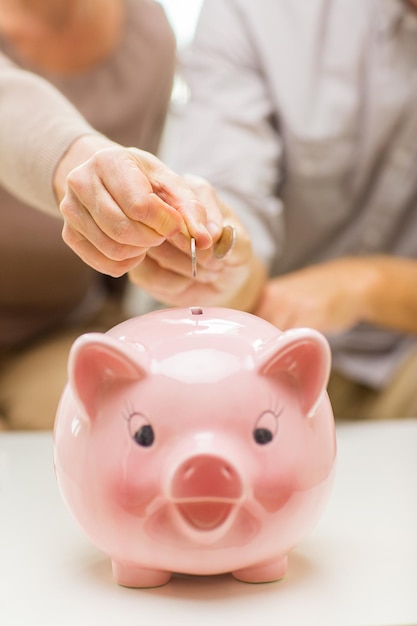 concepto de familia, ahorro, edad y personas - cierre de las manos de la pareja mayor poniendo dinero en monedas en la alcancía