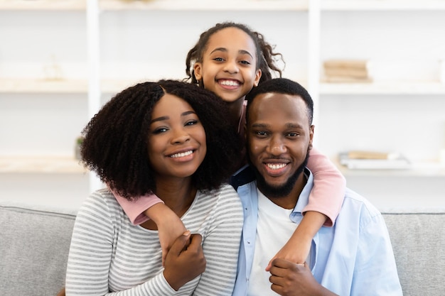 Concepto de familia amorosa feliz. Retrato de una emocionada niña afroamericana abrazando a su mamá y papá desde atrás, padres sentados en un sofá en la sala de estar, posando para una foto mirando la cámara en casa