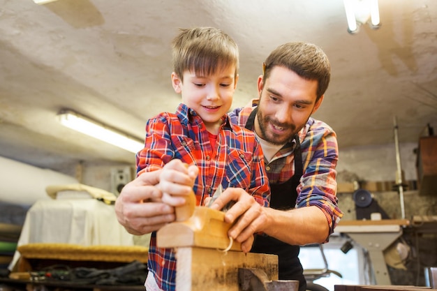 concepto de familia, carpintería, carpintería y personas - padre e hijo pequeño con avión trabajando con tablones de madera en el taller